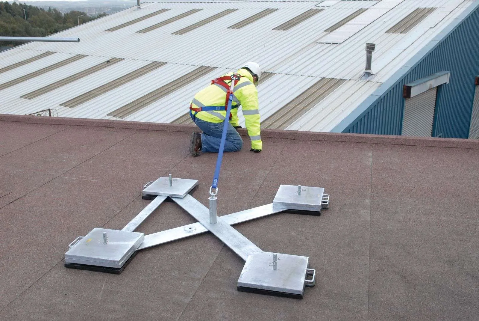 Worker wearing fall protection harness attached to portable roof anchor on industrial rooftop.